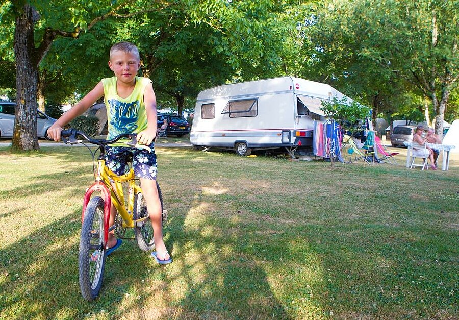 Kind op de fiets voor een caravan op een schaduwrijke staanplaats, zomerse familiesfeer.