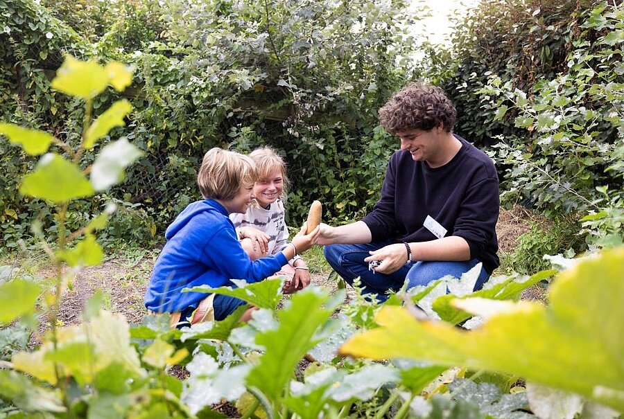 Twee kinderen ontdekken een groente uit de moestuin samen met een begeleider op een ecolabel-camping, een voorbeeld van milieubewust toerisme.