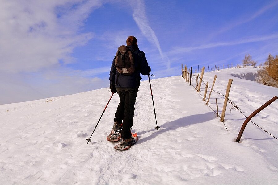 Een sneeuwschoenwandelaar trekt over een besneeuwd pad onder een blauwe hemel, midden in een ongerepte natuur. Geniet van een verblijf op een caravaneige-camping voor een onvergetelijke wintervakantie met Sites et Paysages.