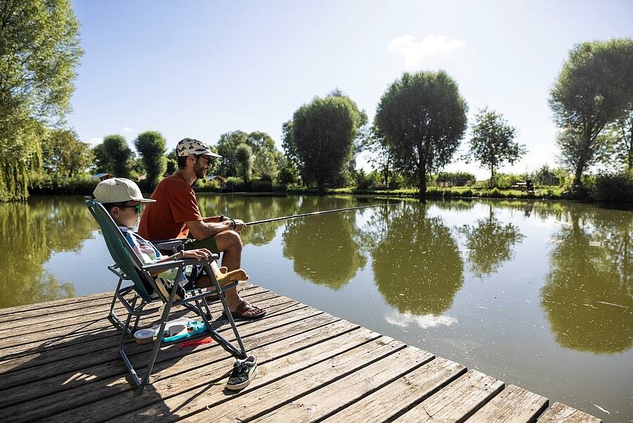 Vader en kind vissen vanaf een houten steiger aan een vijver op een camping vissen, rustige zonnige sfeer.