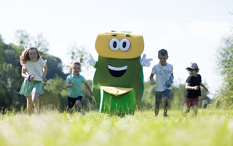 Kinderen rennen door een groen veld samen met PAKO, een vrolijke mascotte, tijdens een familie-animatie op een charme camping vlak bij Honfleur, in Normandië.