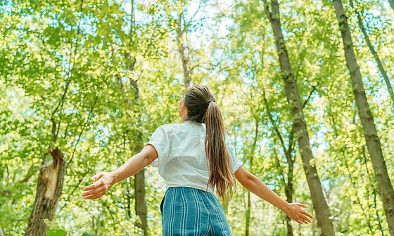 [Translate to Néerlandais:] Jeune fille les bras ouverts en pleine forêt, symbole de reconnexion à la nature et de tourisme durable avec Sites et Paysages.