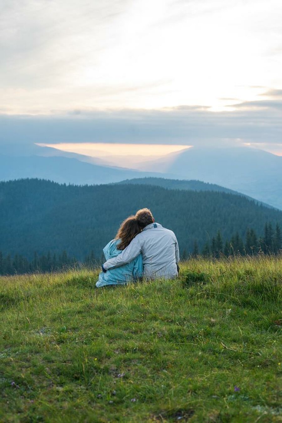 Koppel zit samen in het gras met uitzicht op bergen bij zonsondergang, rustige plek voor een romantisch weekend op de camping.