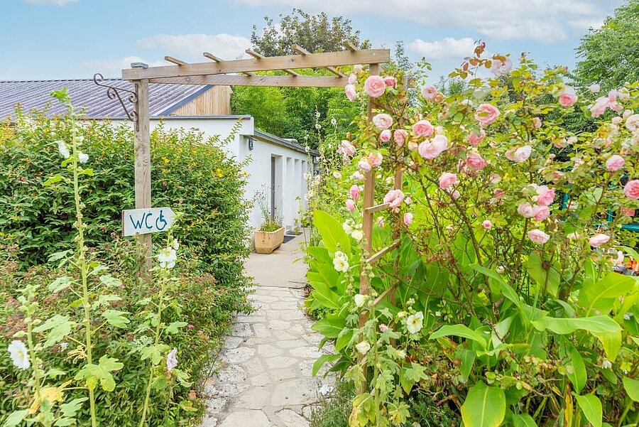 Bloemrijke toegang tot de toegankelijke sanitaire voorzieningen vlak bij een charme camping staanplaats voor caravan, in een verzorgde en natuurlijke omgeving.