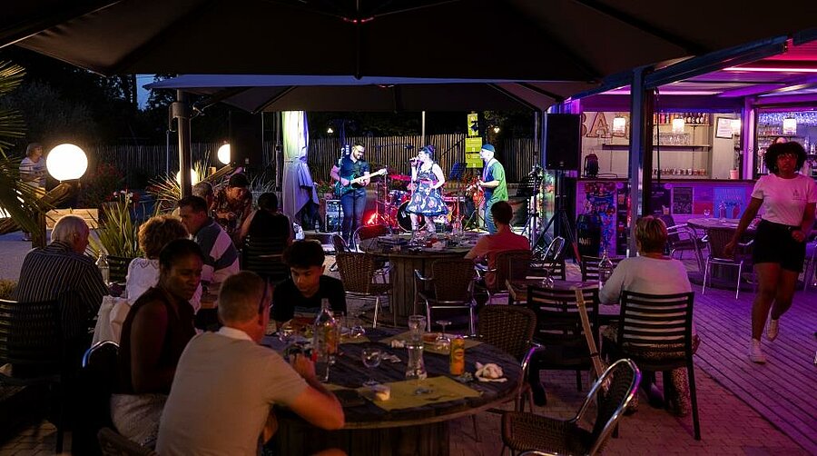 Gezellige campingavond met openluchtconcert, families en vrienden samen aan tafel in een warme sfeer.