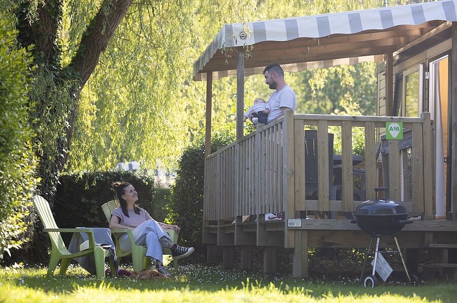 [Translate to Néerlandais:] Famille profitant d’un moment de détente sur la terrasse d’un mobil-home ombragé, dans un camping avec parc aquatique et cadre naturel verdoyant