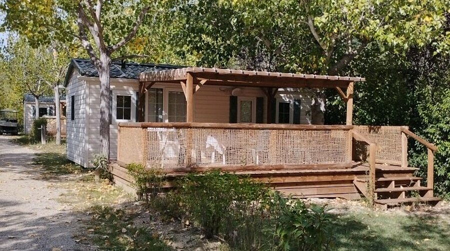 Stacaravan met overdekt houten terras en tuinmeubilair, gelegen tussen de bomen op camping Domaine de l’Écluse bij Mont Ventoux.