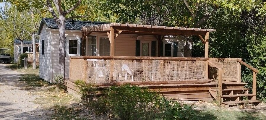 Stacaravan met overdekt houten terras en tuinmeubilair, gelegen tussen de bomen op camping Domaine de l’Écluse bij Nyons.