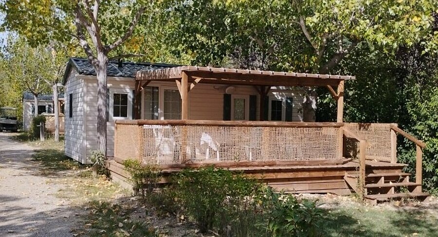 Stacaravan met overdekt houten terras en tuinmeubilair, gelegen tussen de bomen op camping Domaine de l’Écluse in de Drôme.