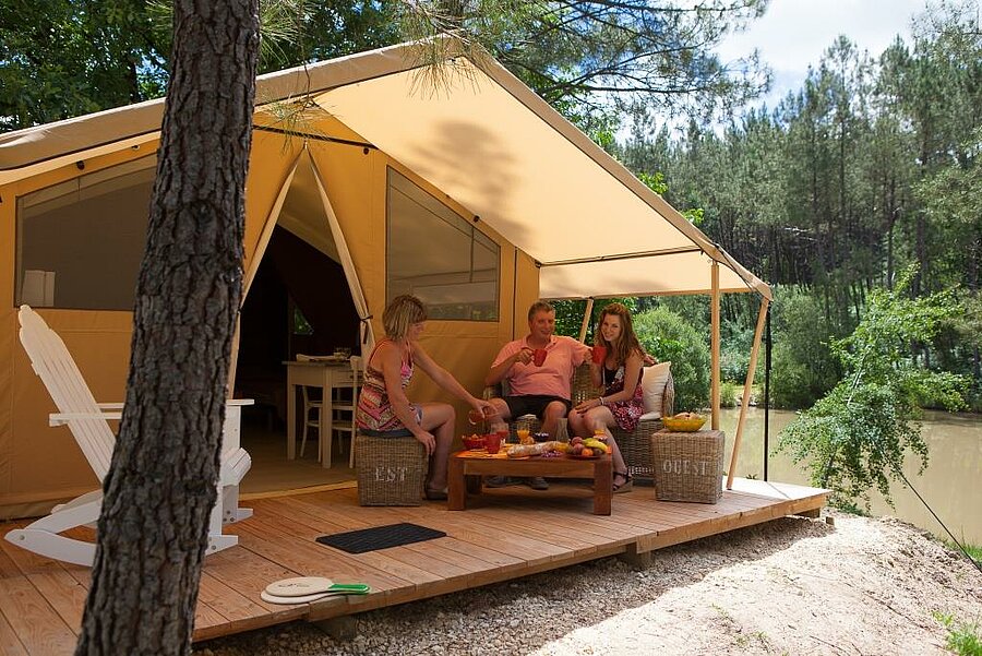 Gezin geniet van een houten terras voor een lodgetent op een campsite in Nouvelle-Aquitaine, aan het meer en omgeven door dennenbomen.