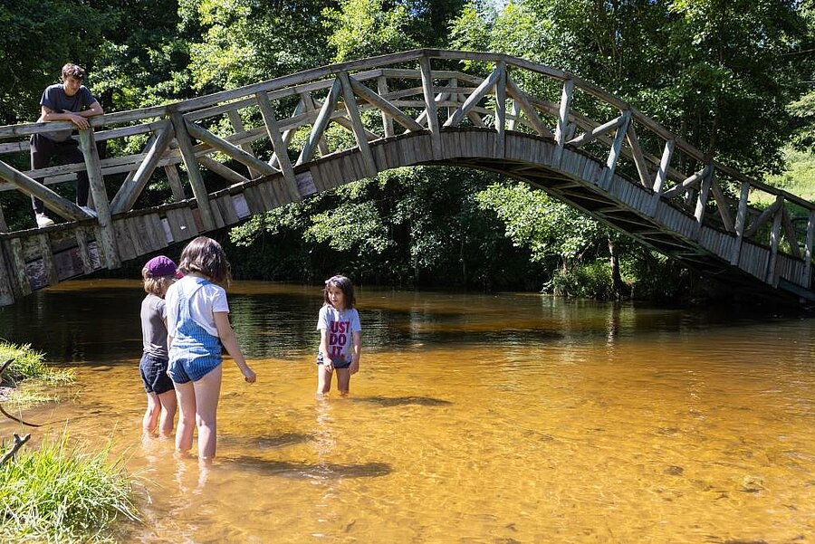[Translate to Néerlandais:] Enfants jouant dans une rivière peu profonde sous un pont en bois, dans un camping pas cher et nature idéal pour des vacances familiales au bord de l'eau
