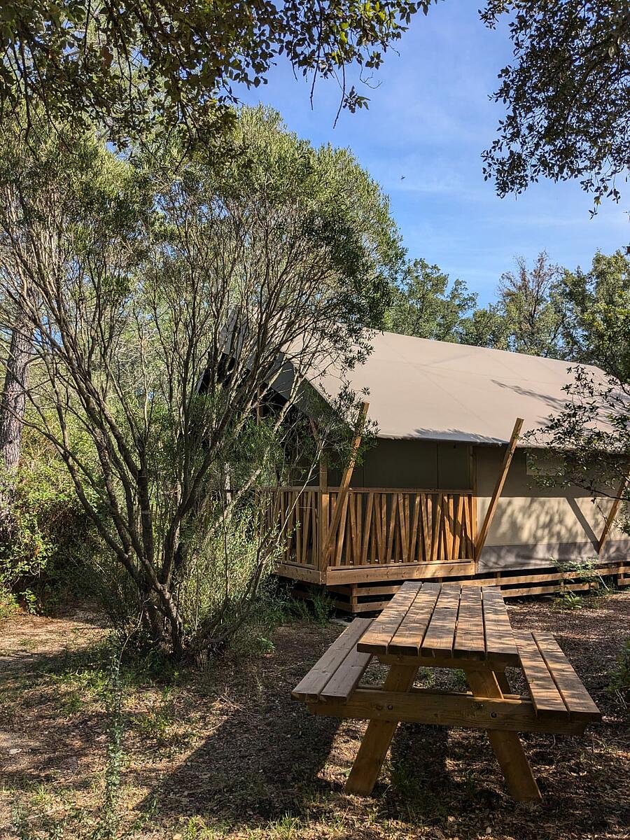 Een houten en canvas lodge-tent met terras, omgeven door groen en uitgerust met een picknicktafel op een camping in Corsica, ideaal voor een comfortabel verblijf midden in de natuur.