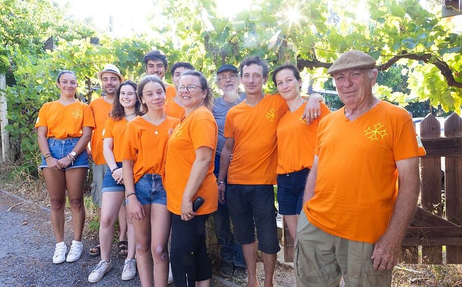Glimlachend team van camping La Pinède bij Carcassonne in oranje shirts onder een pergola met wijnranken.