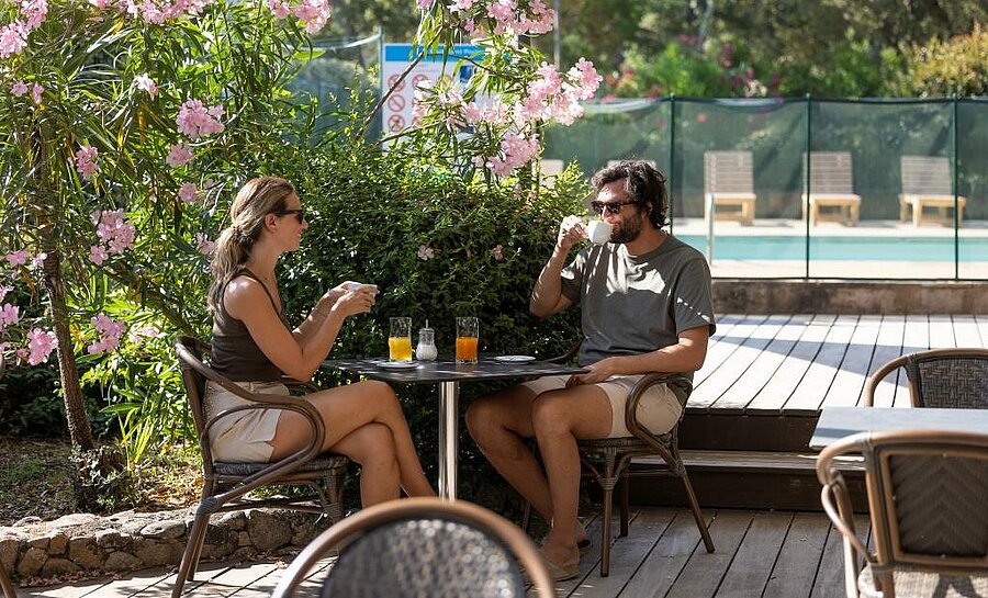 Koppel op schaduwrijk terras met drankjes naast het zwembad van een camping op Corsica