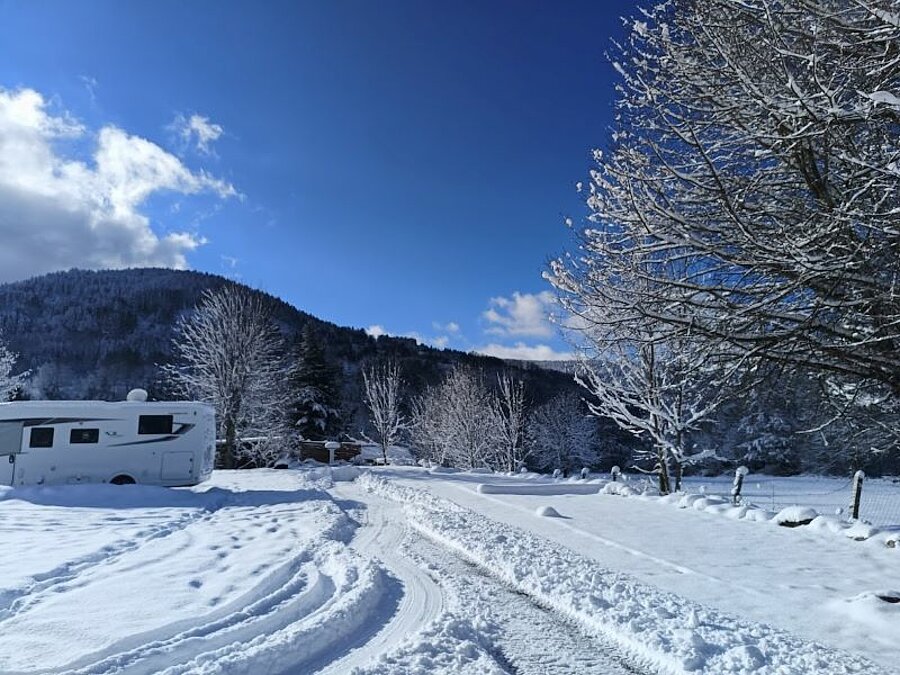 Caravaneige camping onder de sneeuw met een geparkeerde camper in een winters berglandschap, omringd door besneeuwde bomen. Een ideale setting voor een winterse buitenvakantie.
