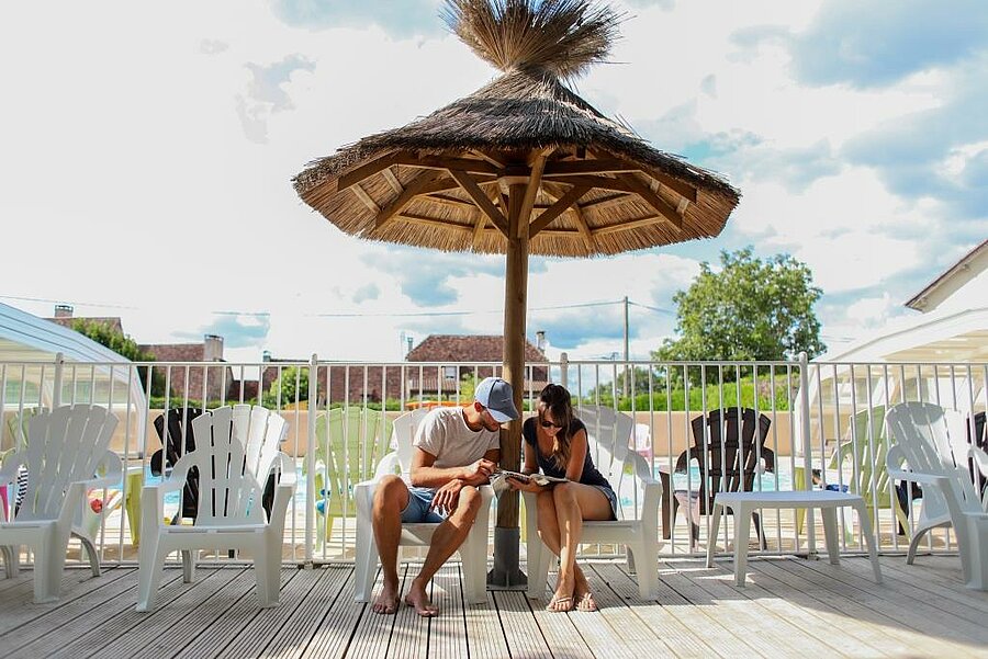 [Translate to Néerlandais:] Jeune couple en pleine détente sous un parasol en bord de piscine dans un camping pas cher, à la recherche du meilleur rapport qualité-prix.
