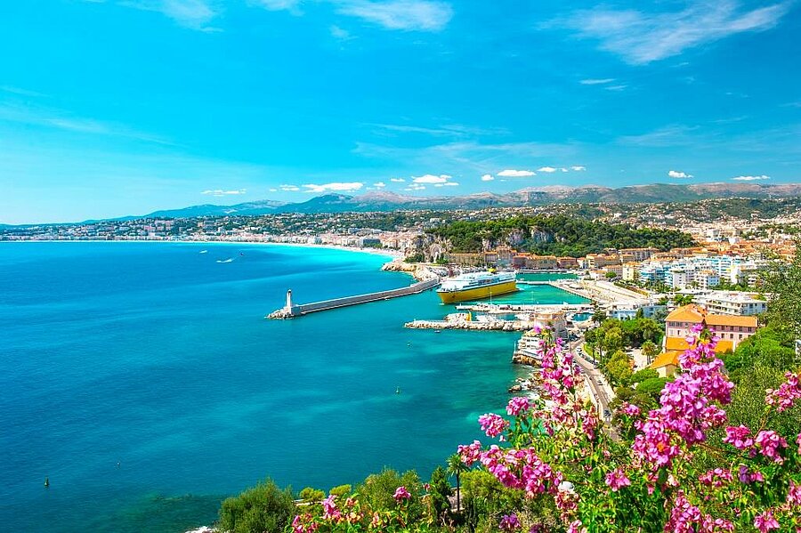 Uitzicht op de haven van Nice, de turquoise Middellandse Zee en omliggende heuvels met mediterrane bloemen, vlak bij een camping aan de Côte d’Azur.
