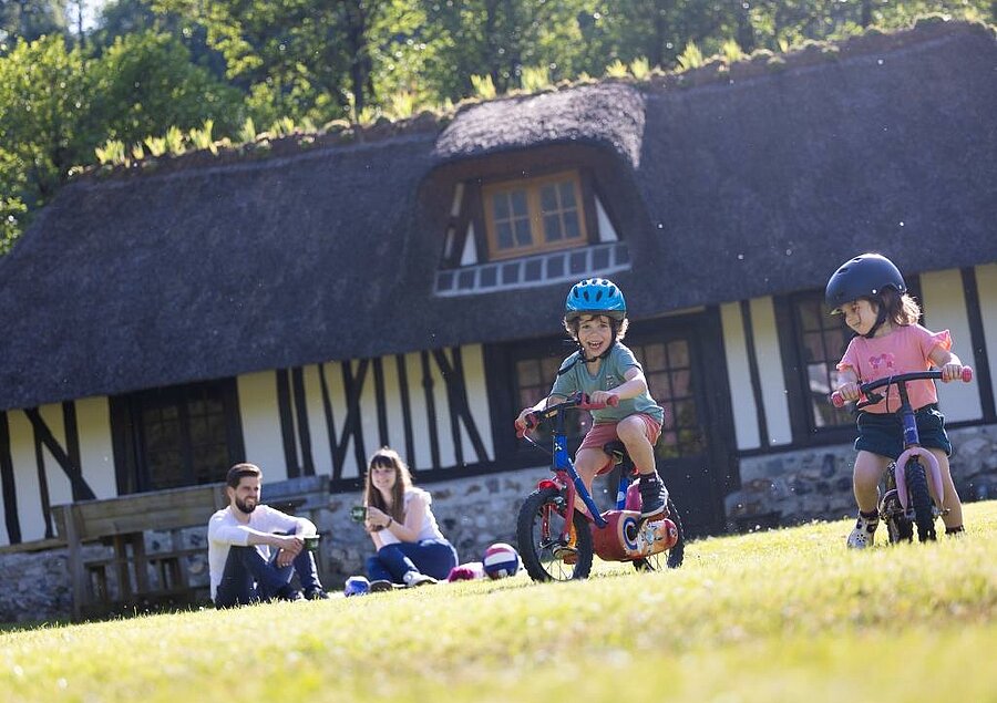 Twee kinderen fietsen voor een typische Normandische accommodatie, terwijl hun ouders ontspannen op een charme camping vlak bij Honfleur.