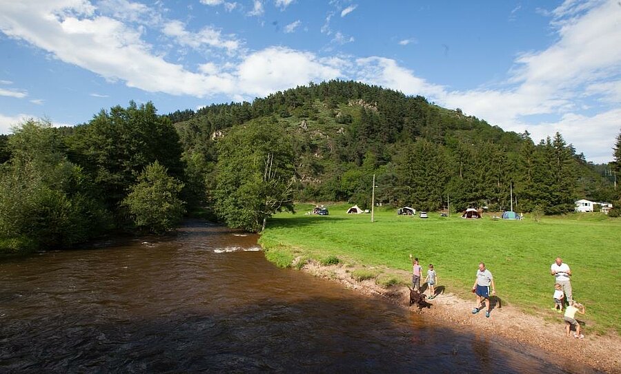 Gezin wandelt langs een rivier bij een camping aan het meer, groene staanplaatsen en bosrijke heuvels op de achtergrond.